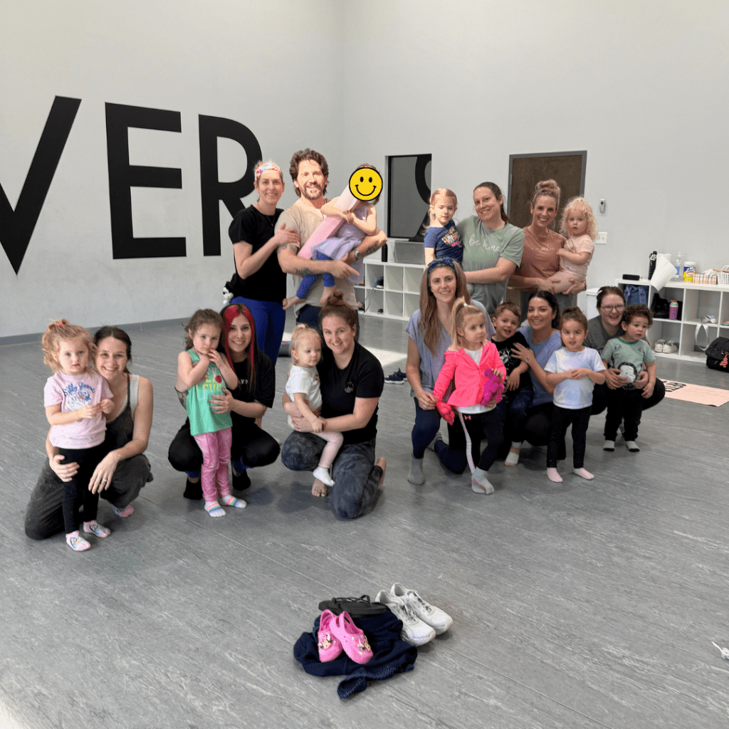 Group of children and adults during a toddler yoga class in a spacious studio, with a playful atmosphere and yoga props in the background.